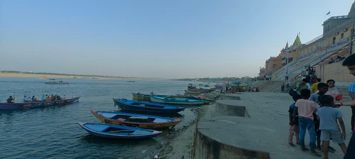 Boats moored along the Ganga ghats at dusk, with visitors on the steps